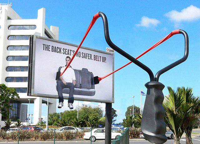 billboard with large slingshot telling backseat passengers to buckle up