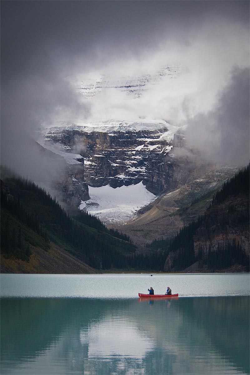 break in the clouds as two people in canoe see a spec of sun hiting the victoria glacier above a glacial lake in lake louise banff alberta canada