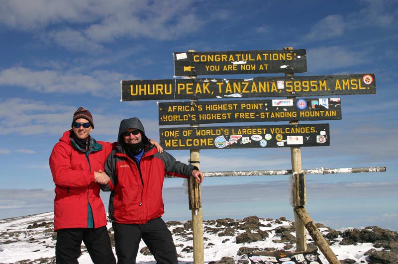 two climbers in front of sign at the top of mount kilimanjaro the highest point in africa