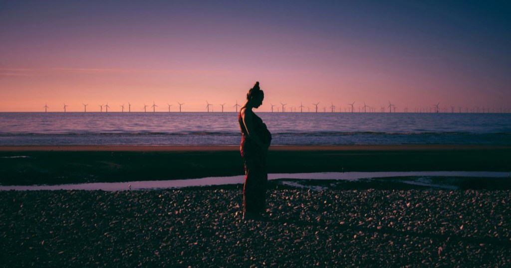 A pregnant woman on the beach at sunset with wind turbines in the distance