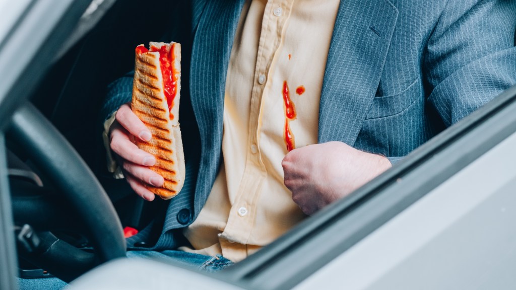 A man with a messy sandwich has spilled sauce on his shirt while sitting in a car