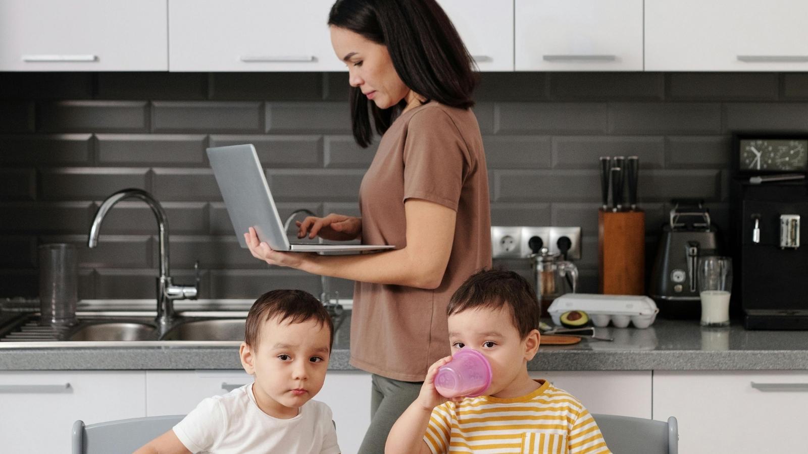 A mother working on her laptop while her children eat breakfast at home.