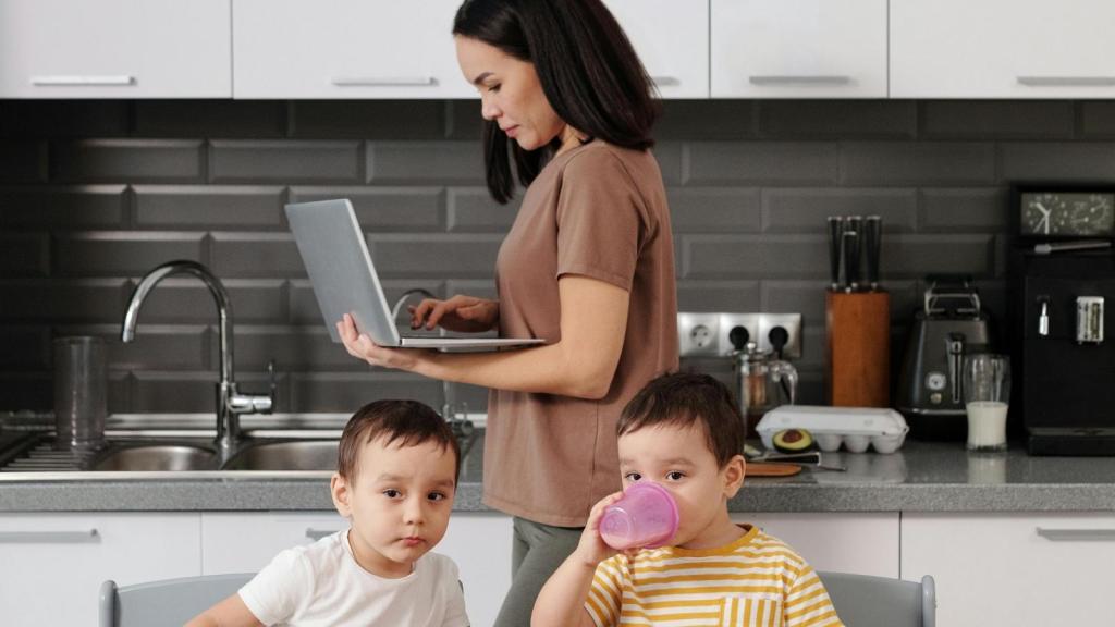 A mother working on her laptop while her children eat breakfast at home.