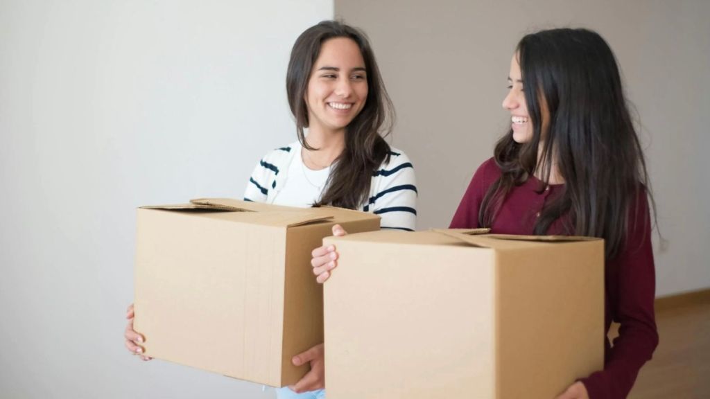 two smiling young woman carrying moving boxes