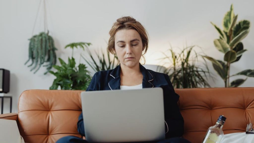 Woman looking at a laptop wehile sitting on the couch with a bottle of alcohol beside her