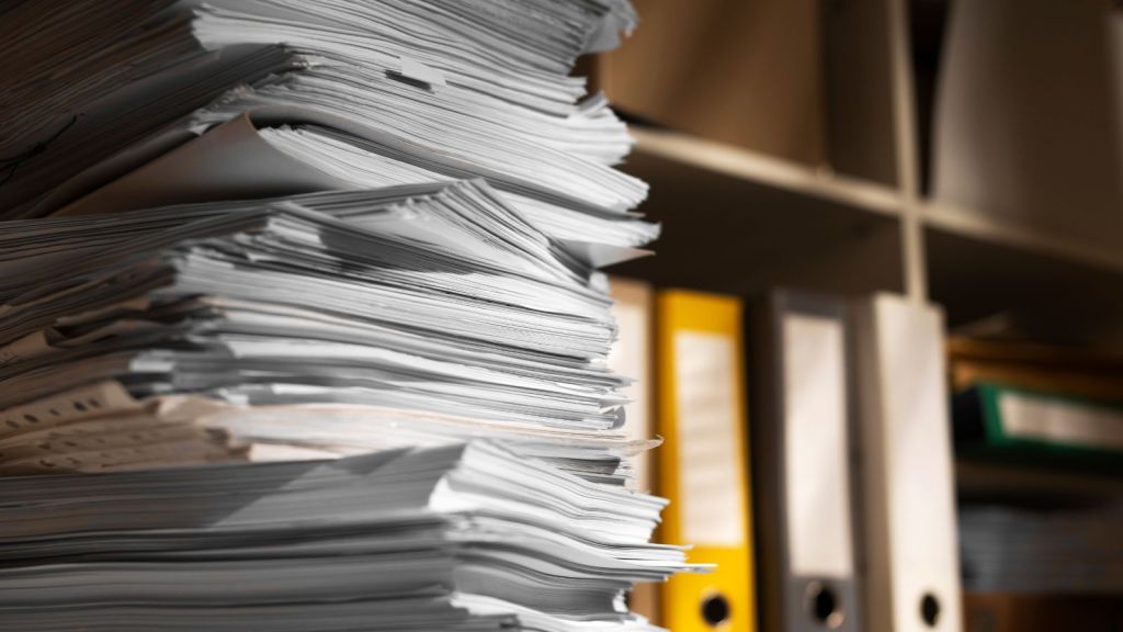 A stack of papers on the table and some file folders at the shelf
