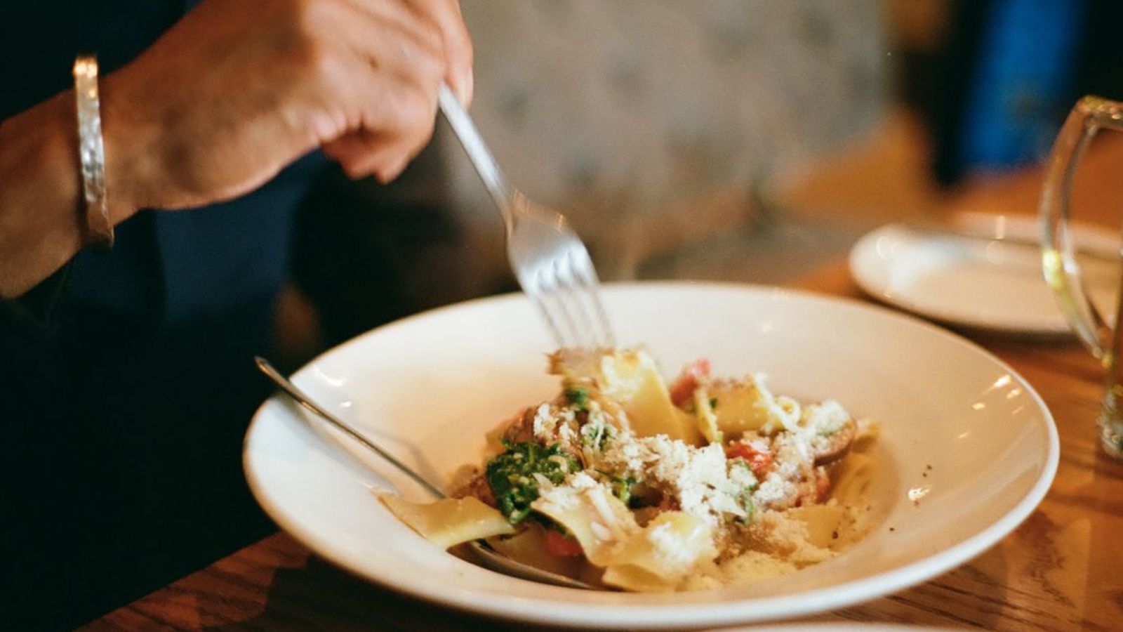 Man eating a bowl of pappardelle pasta topped with parmesan and vegetables at a fancy restaurant