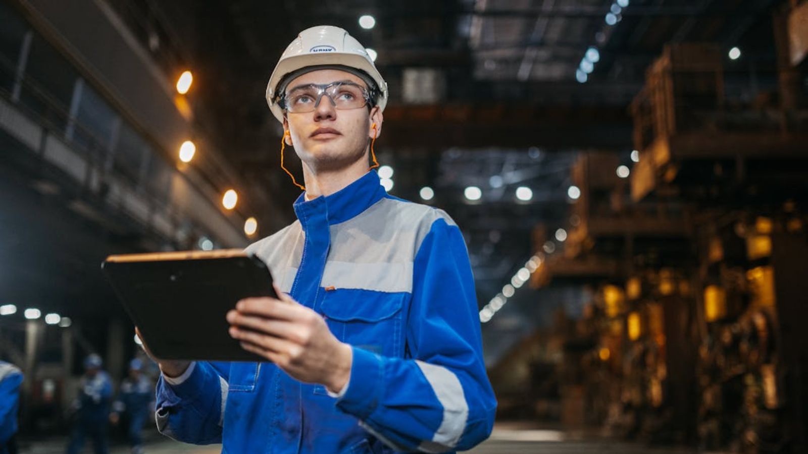 An engineer in safety gear using a tablet in a large industrial factory setting
