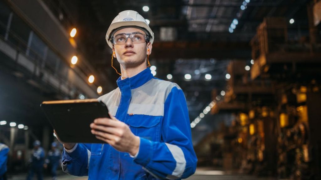 An engineer in safety gear using a tablet in a large industrial factory setting