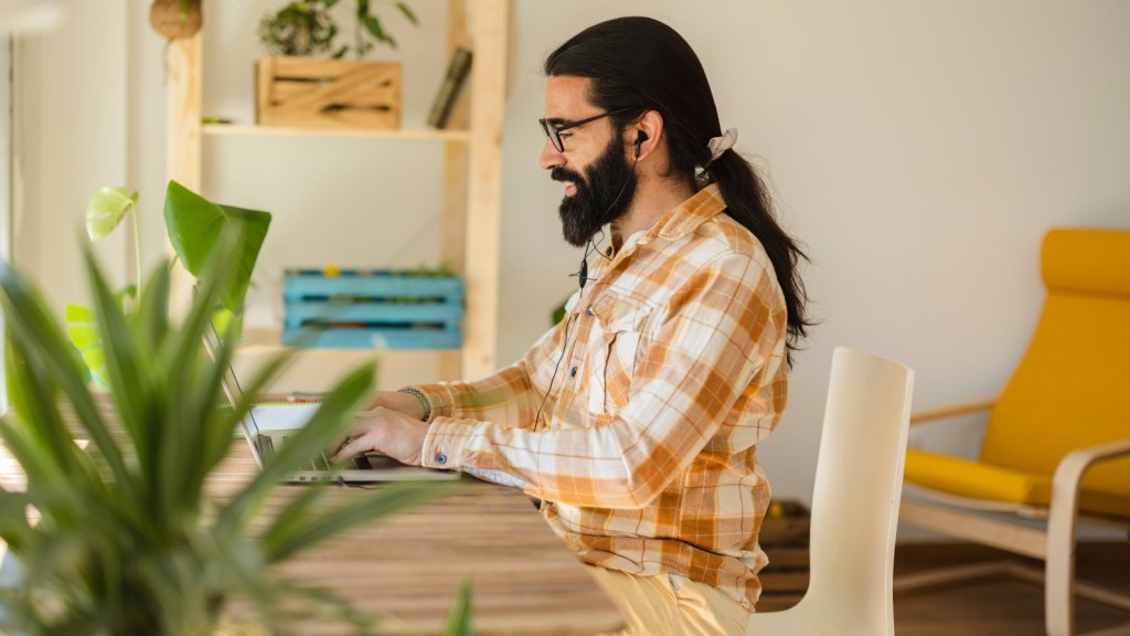 A long haired man sitting at a desk with a laptop