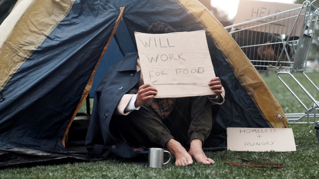 A man in a tent holds a sign that reads "will work for food"