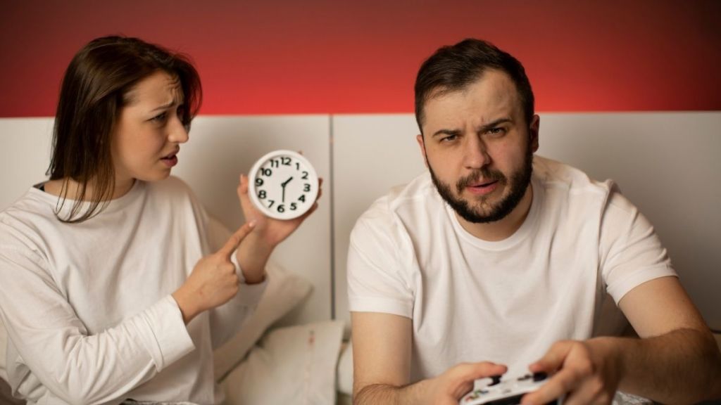 man playing video games while his wife points out the time