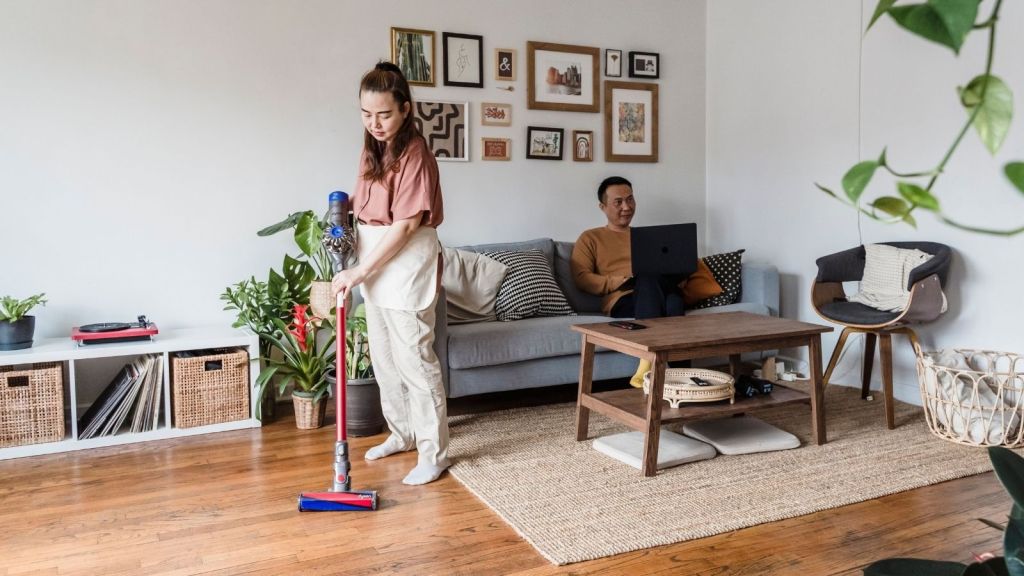 A woman cleaning while a man sits on the couch