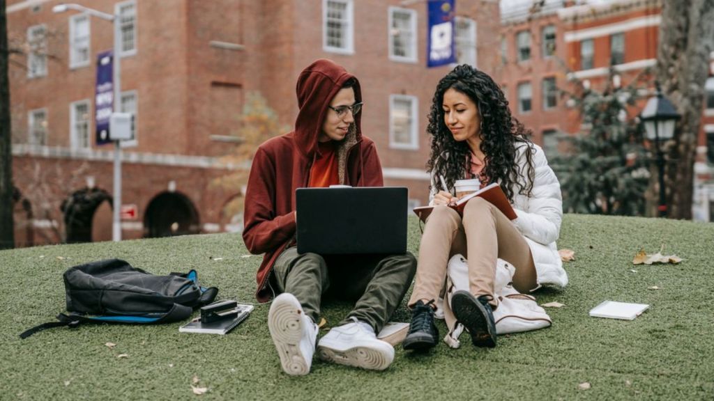 Two college students study together on campus lawn with laptop and notes