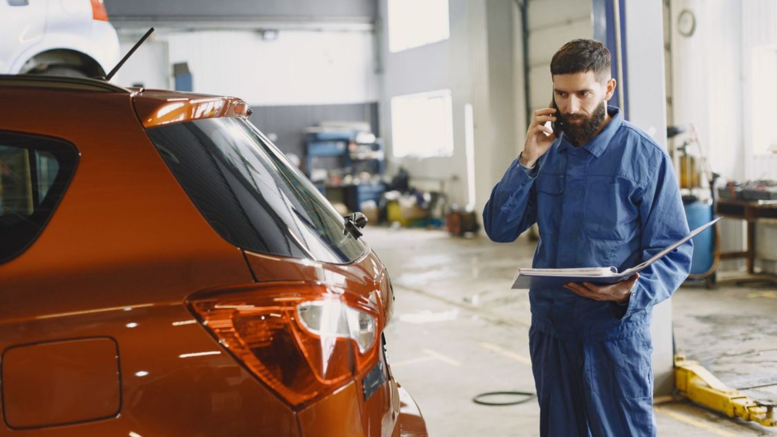 Mechanic in a blue uniform talks on phone beside a red car in an auto repair shop