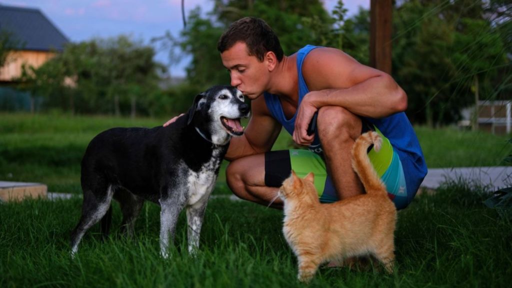Man crouches in a garden kissing his dog while a cat looks on