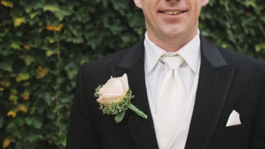 A groom in a classic black tuxedo with a white boutonniere stands against green foliage
