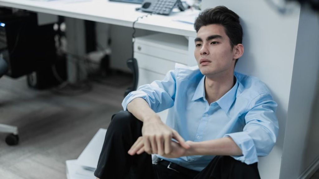 exhausted Asian man sits on an office floor looking reflective and stressed, surrounded by paperwork