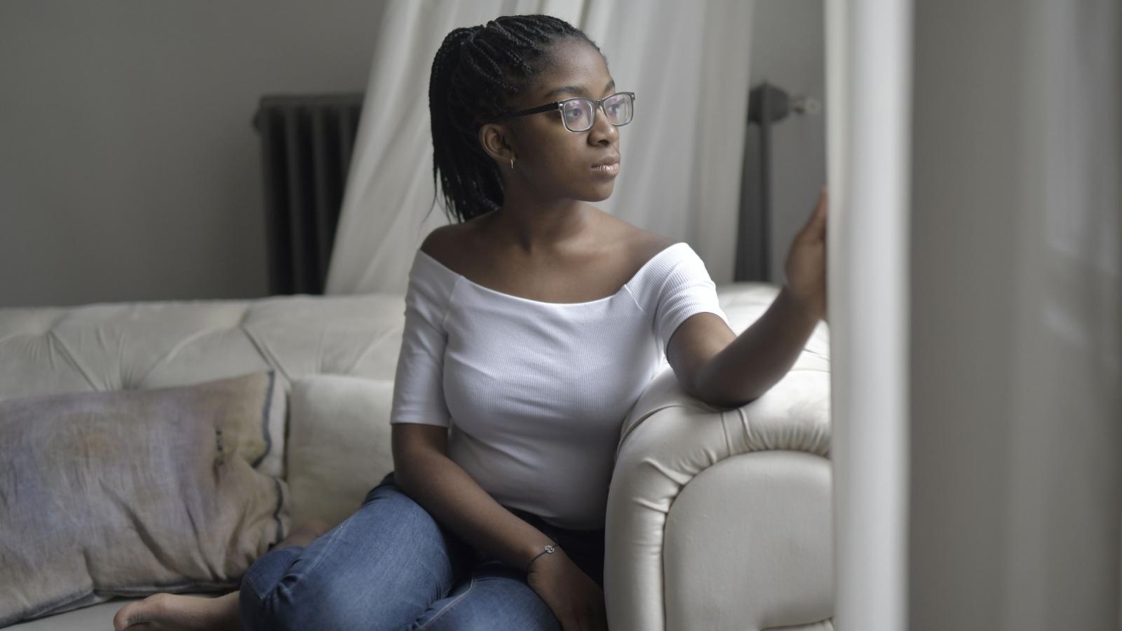 A Renter Is Living in Total Fear After a Downstairs Neighbor Weaponized a Broom Handle Woman looking out the window at her apartment