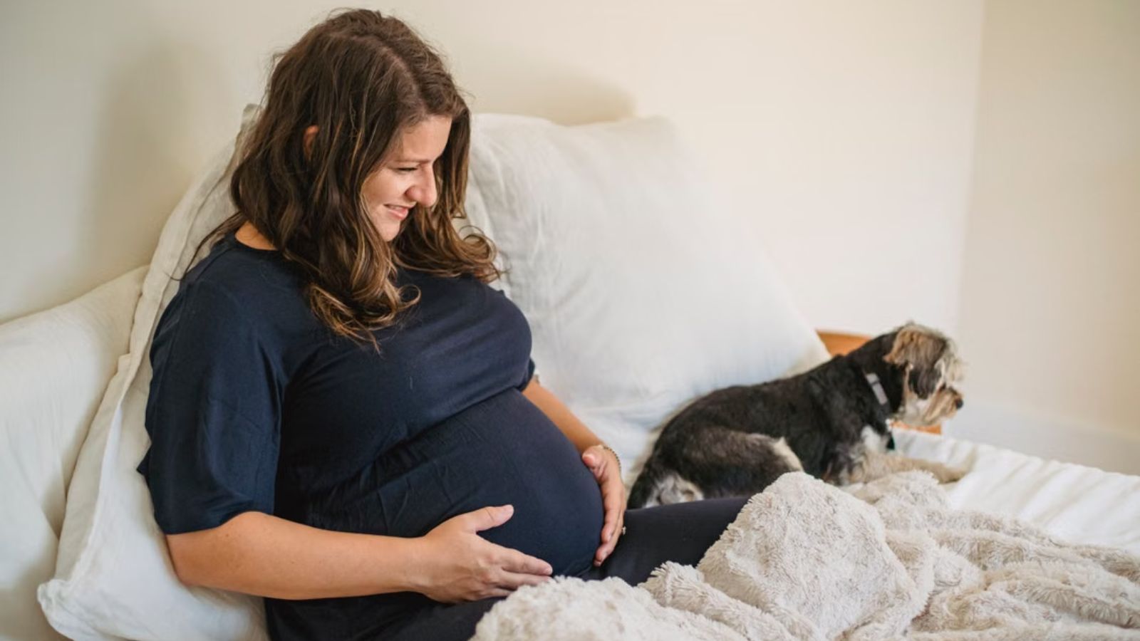 Shes Pregnant and Struggling With Their Difficult Dog, But Her Husband Refuses To Entertain The Idea Pregnant woman touching her belly and a dog sitting beside her on the bed