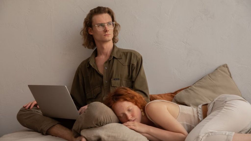 Man using a laptop on bed while woman sleeps beside him