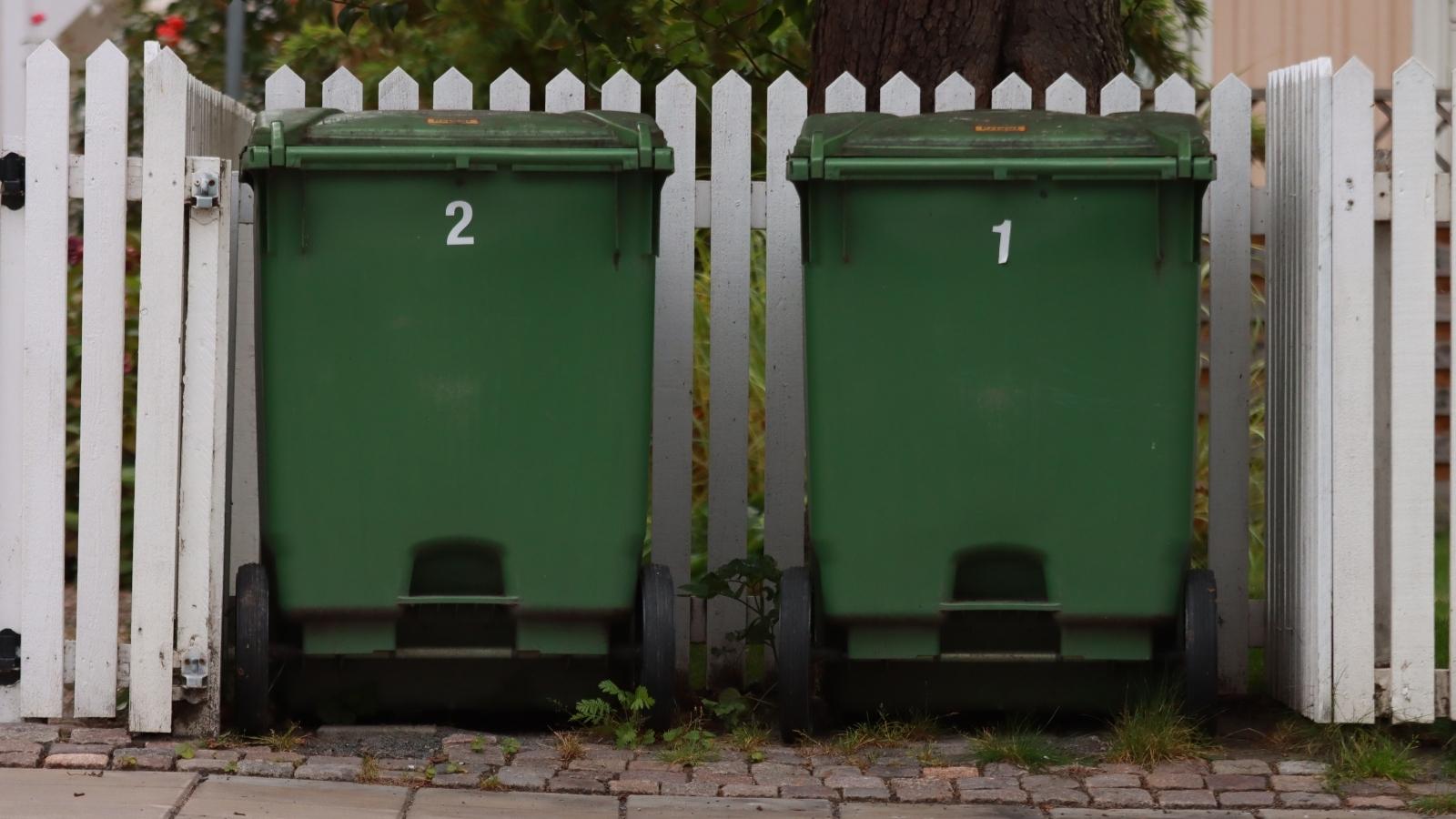 Neighborhood Feud Escalates After Neighbor Intentionally Blocks Parking Spaces Over Trash Can Boundary Two green trash cans parked outside a white picket fence