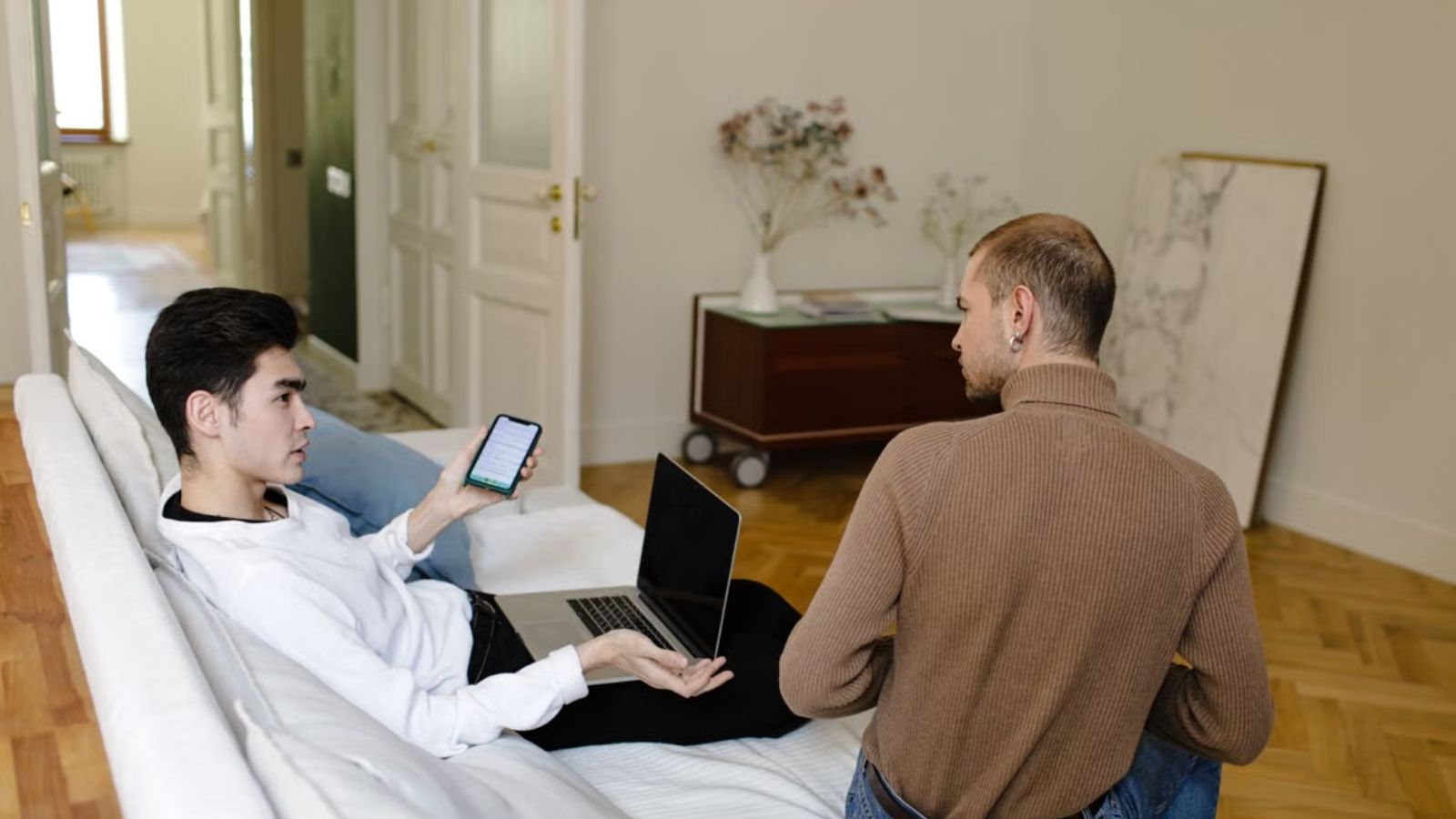 He Paid Every Bill While His Roommate Recovered—Now He’s Being Called Cold for Asking to Split Rent Again Two men engaging in a conversation at home while sitting on the couch