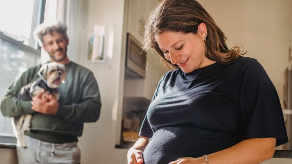 A joyful pregnant woman and her husband smile at each other while holding their pet dog indoors