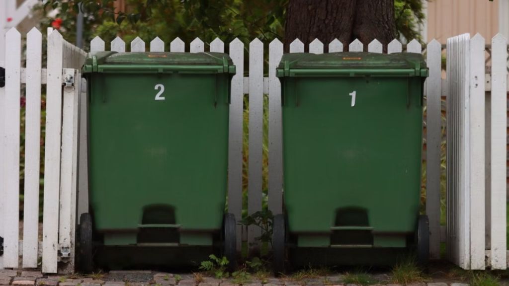 Two green trash bins beside a white picket fence