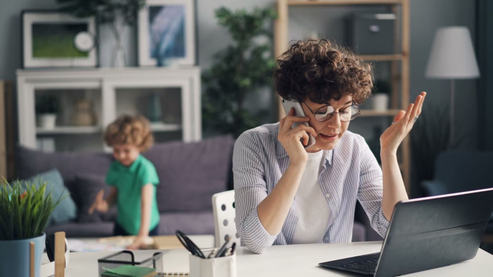 Woman is talking to the phone in front of a tablet while a child is playing at the back