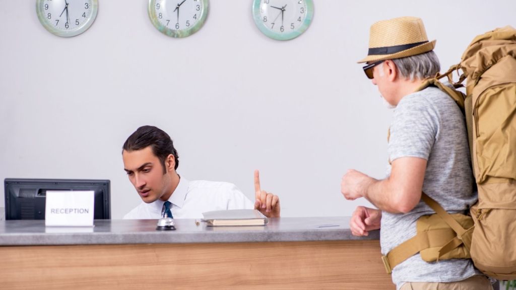 old man wearing a large backpack talking to hotel front desk worker
