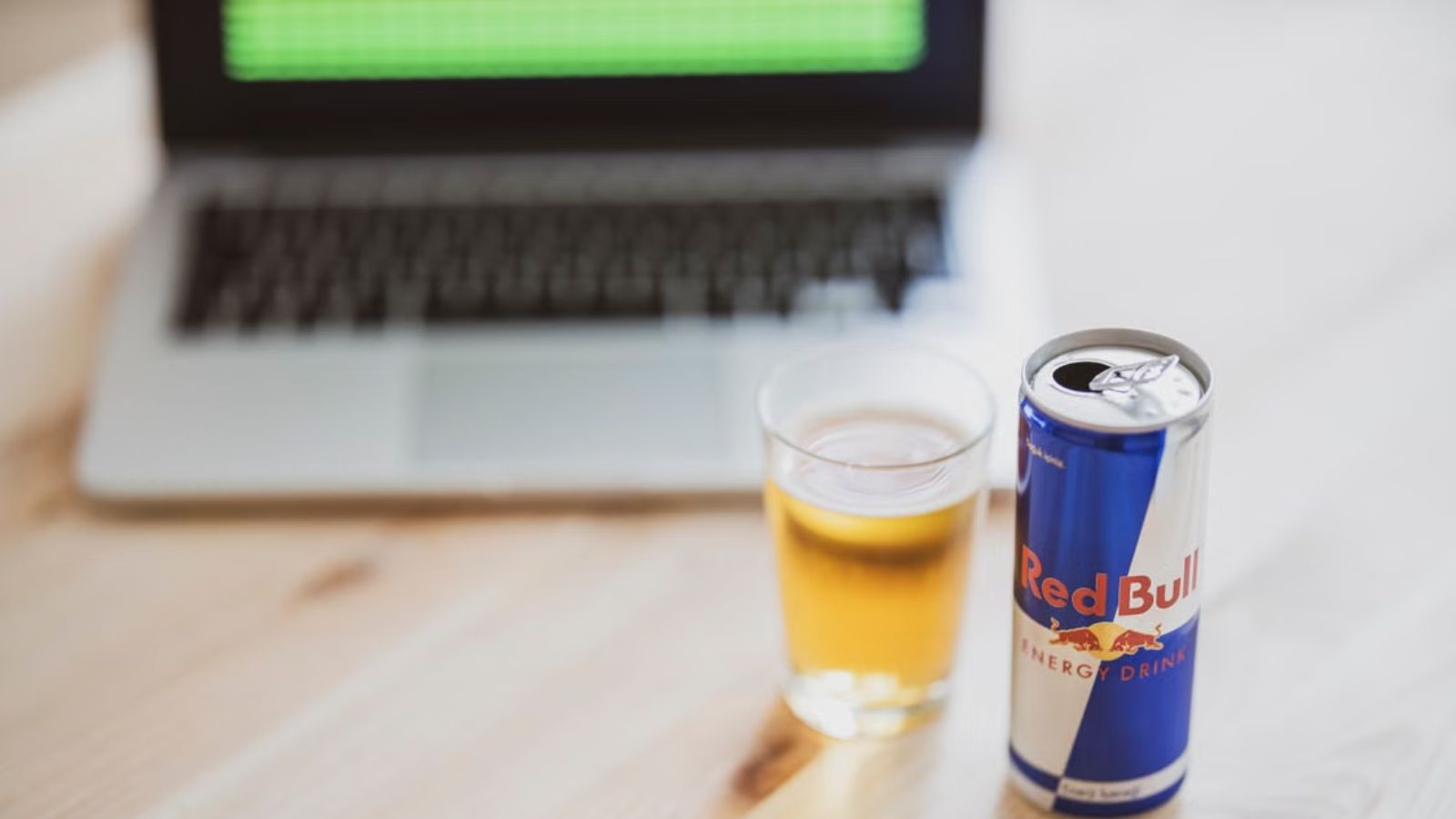 She Drank an Unclaimed Red Bull Sitting in the Fridge for Weeks—Now a Coworker Is Threatening to Report Her to HR for Grand Theft A can of Red Bull and a glass on a wooden desk with a laptop at the back