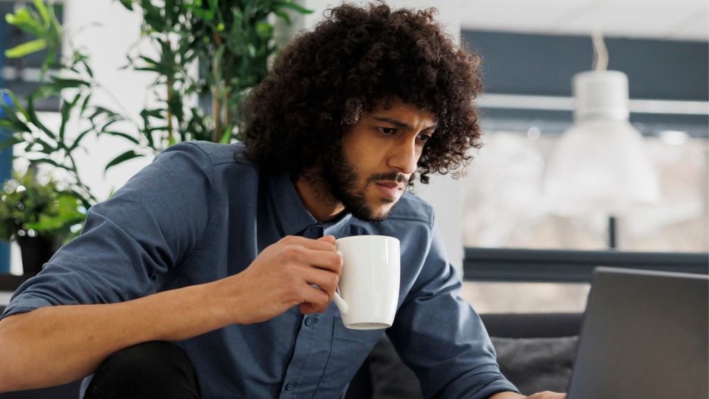 Man holding a cup with one hand and intently looking at his laptop