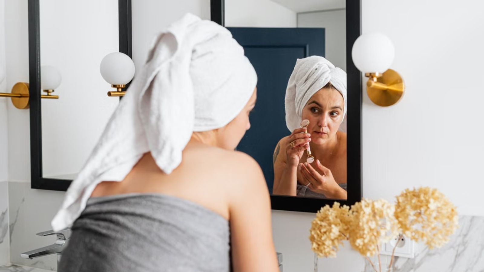 She Hosted Her Friend for a Two Week Reset—Then Caught Her Wearing Her Designer Clothes and Using $200 Skincare Products Without Asking Woman wrapped in towel is using a face roller in the bathroom