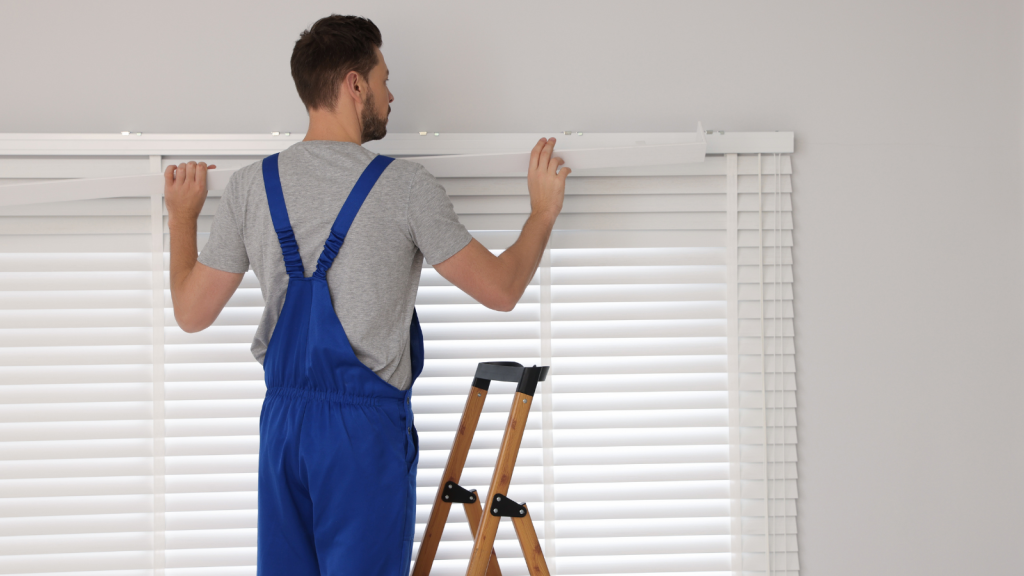 young worker man in uniform installing window blinds on ladder