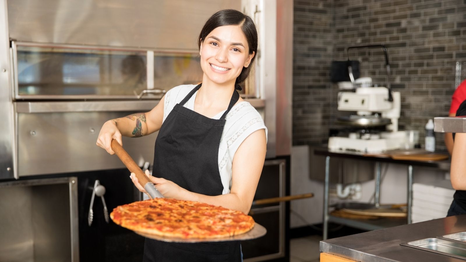 She Stepped Up to Cover Every Last Minute Shift—Only to Be Scolded for Hitting Overtime Hours woman holding a pizza on a pizza peel at a restaurant
