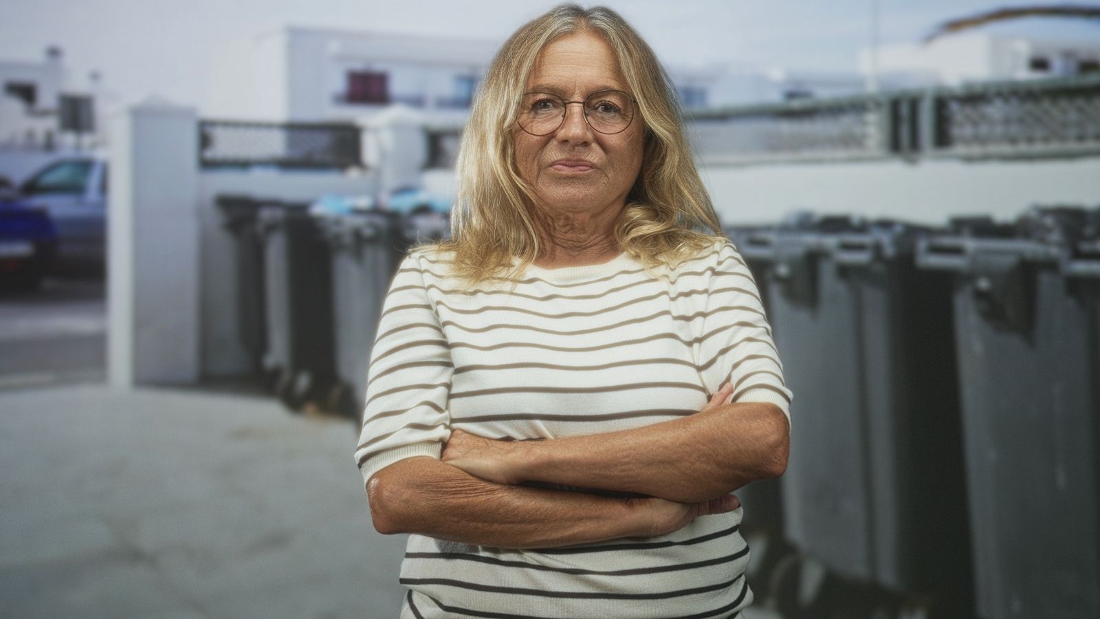 older woman crossing her arms while standing in front of trash bins