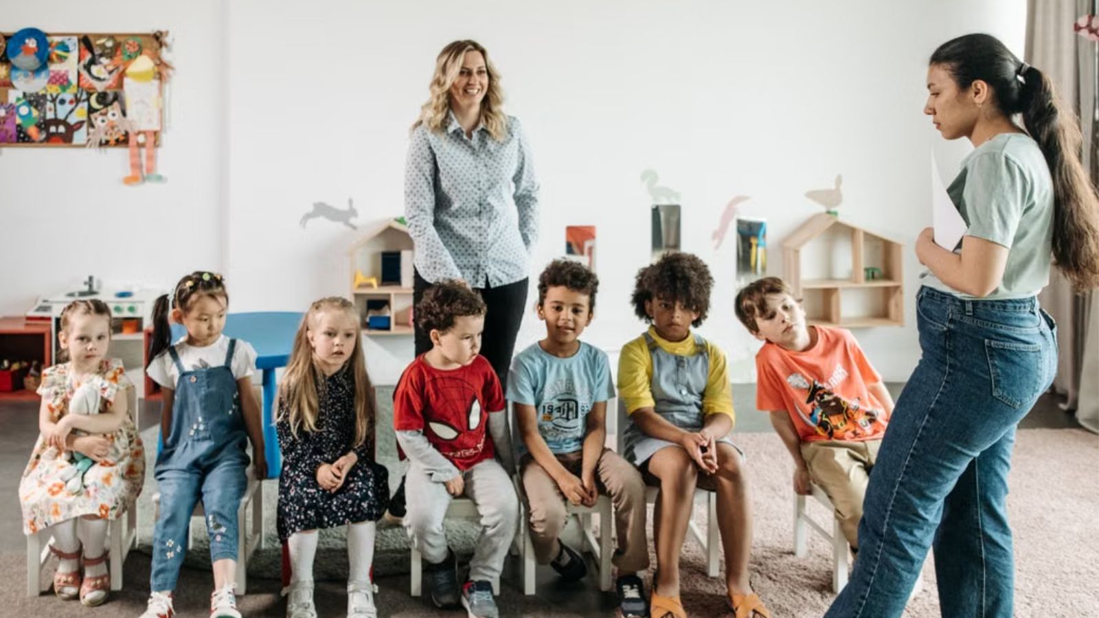Kids sitting on a straight line with teachers standing near them inside the classroom